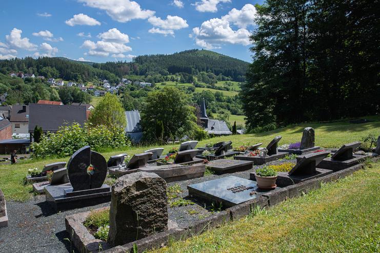 Gräber mit bunten Blumen auf einem Friedhof, umgeben von einer grünen Landschaft und blühendem Himmel.