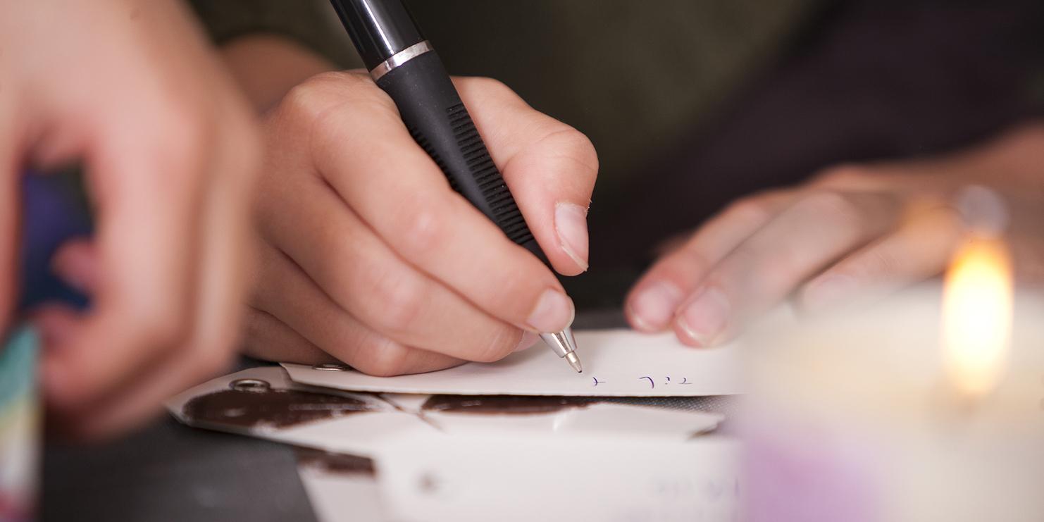 Nahaufnahme einer Hand, die mit einem Kugelschreiber auf einem Zettel schreibt, im Hintergrund eine brennende Kerze.